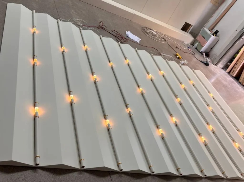 The white zigzag surface has protruding triangular ridges with warm integrated LED linear lights embedded in the V-shaped valleys, paired with a marble reception desk.