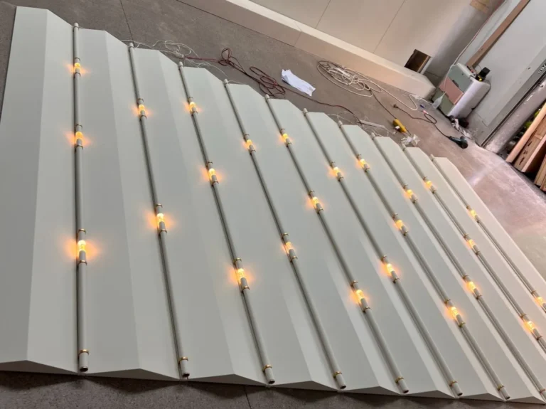 The white zigzag surface has protruding triangular ridges with warm integrated LED linear lights embedded in the V-shaped valleys, paired with a marble reception desk.